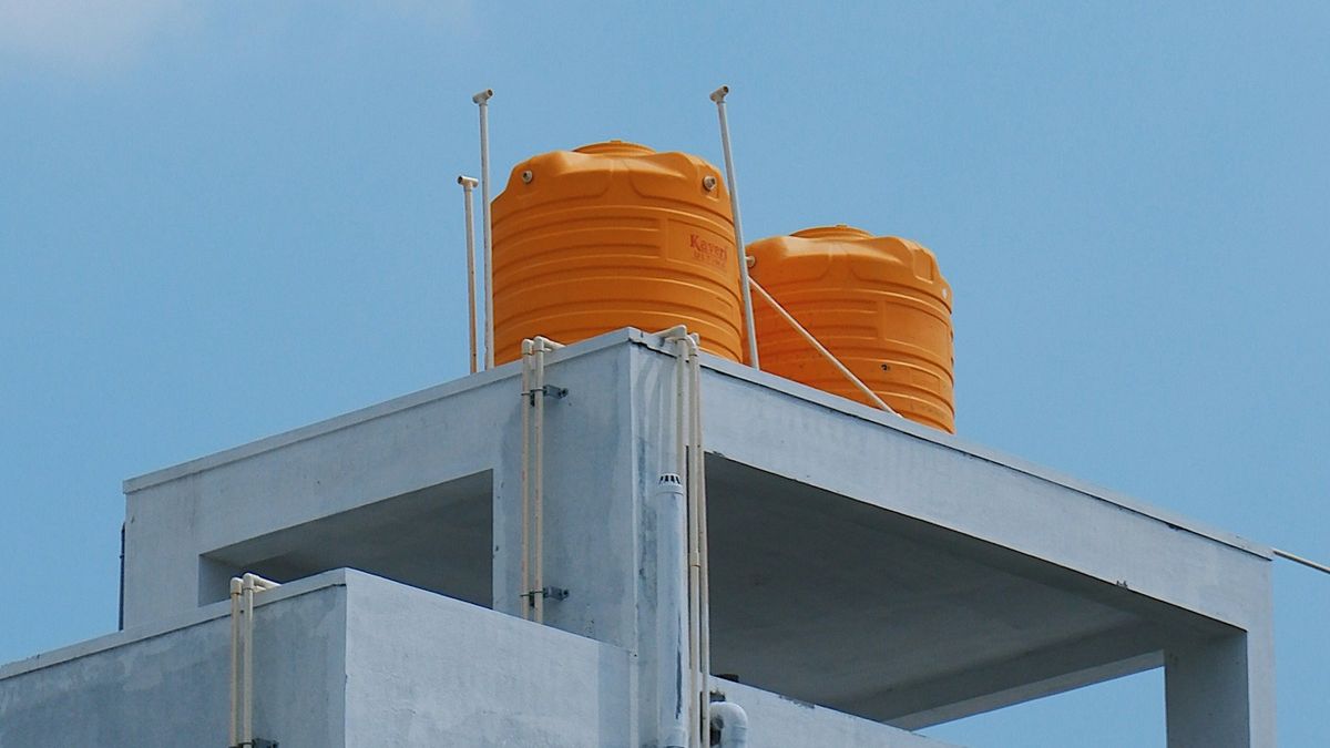 Duas caixas d'água plásticas laranja sobre a laje de cobertura de um prédio, com canos de PVC expostos ao sol e céu azul ao fundo