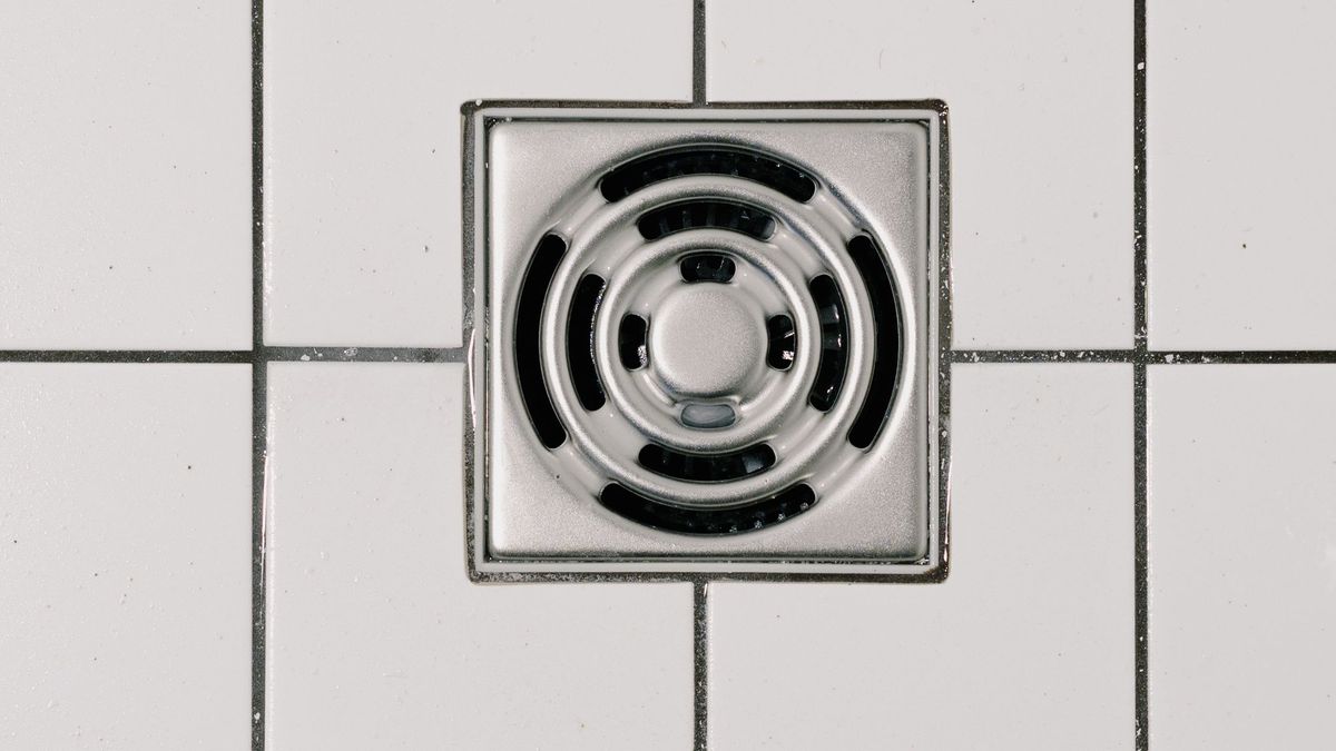 Ralo quadrado de inox com grelha circular instalado no piso de azulejos brancos de um banheiro