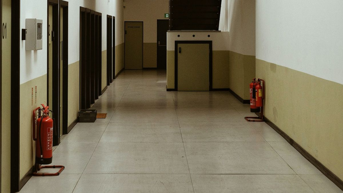 Corredor de edificação com piso claro e paredes bicolores, com extintores de incêndio vermelhos em suportes de chão dos dois lados e placa de saída de emergência ao fundo