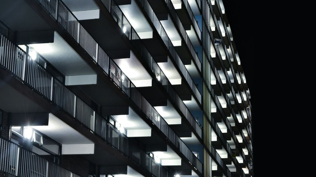 Fachada de prédio residencial fotografada de baixo para cima à noite, com fileiras de varandas iluminadas e guarda-corpos metálicos contra o céu escuro