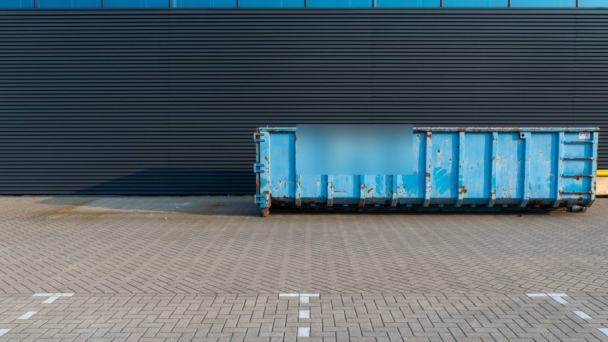 Caçamba de entulho estacionária azul parada em pátio pavimentado junto à parede de um prédio
