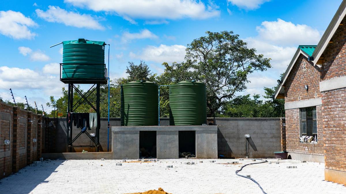 Três reservatórios plásticos verdes de água instalados em um pátio, dois sobre base de concreto e um elevado em torre metálica, com céu azul ao fundo