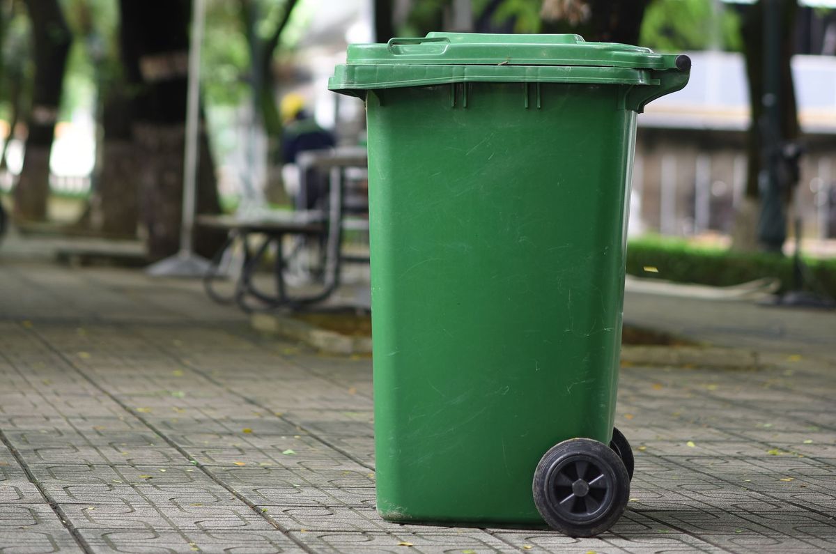 Contentor de lixo plástico verde com tampa e rodízio em área externa pavimentada