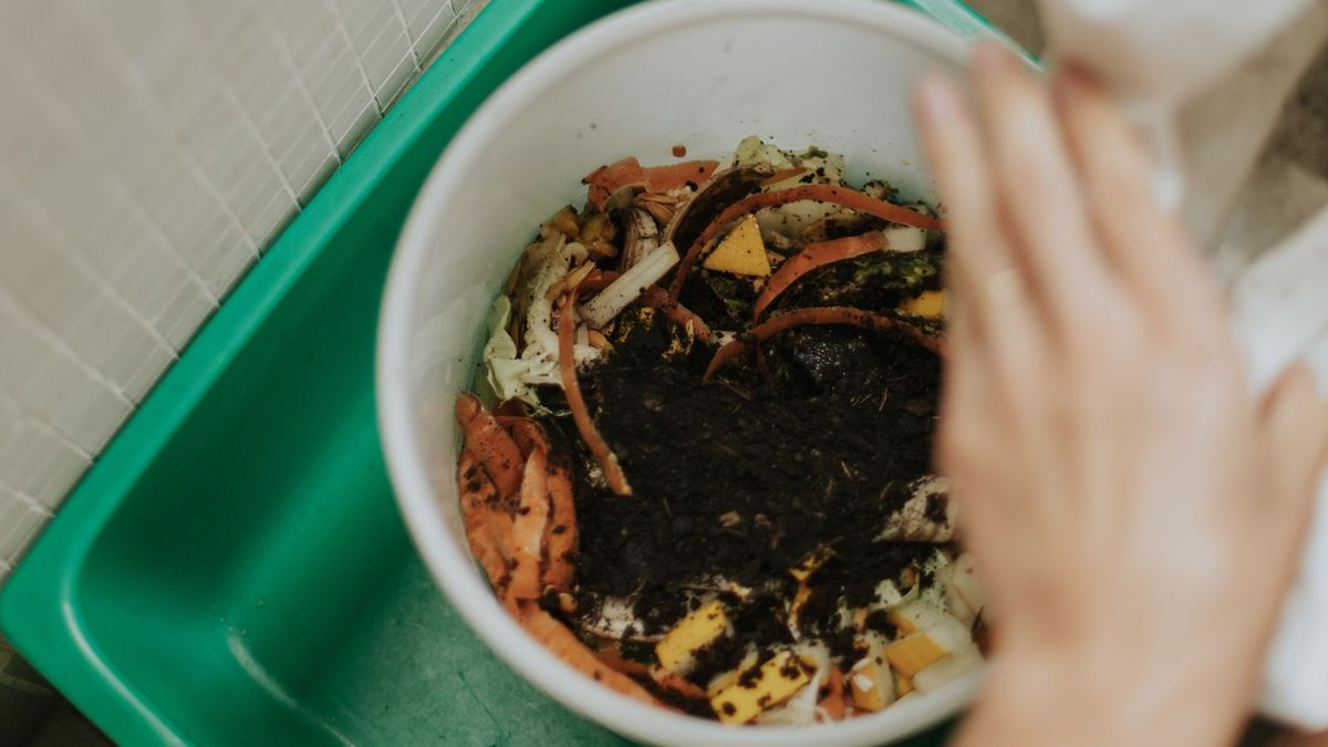 Balde branco com cascas de cenoura, folhas e restos de legumes cobertos por terra escura de composto, sobre uma bacia verde ao lado de uma parede de pastilhas