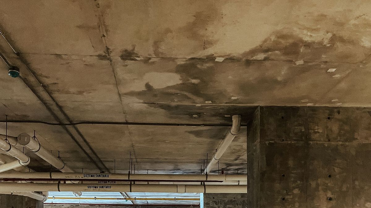 Teto de concreto de uma garagem subterrânea com grandes manchas escuras de umidade, tubulações de esgoto e água pluvial e um pilar ao lado