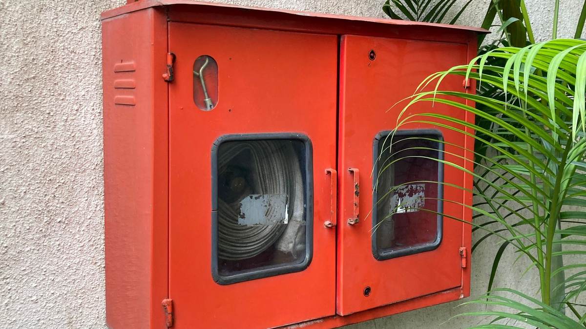 Caixa de mangueira de incêndio vermelha instalada na parede externa de um prédio, ao lado de plantas