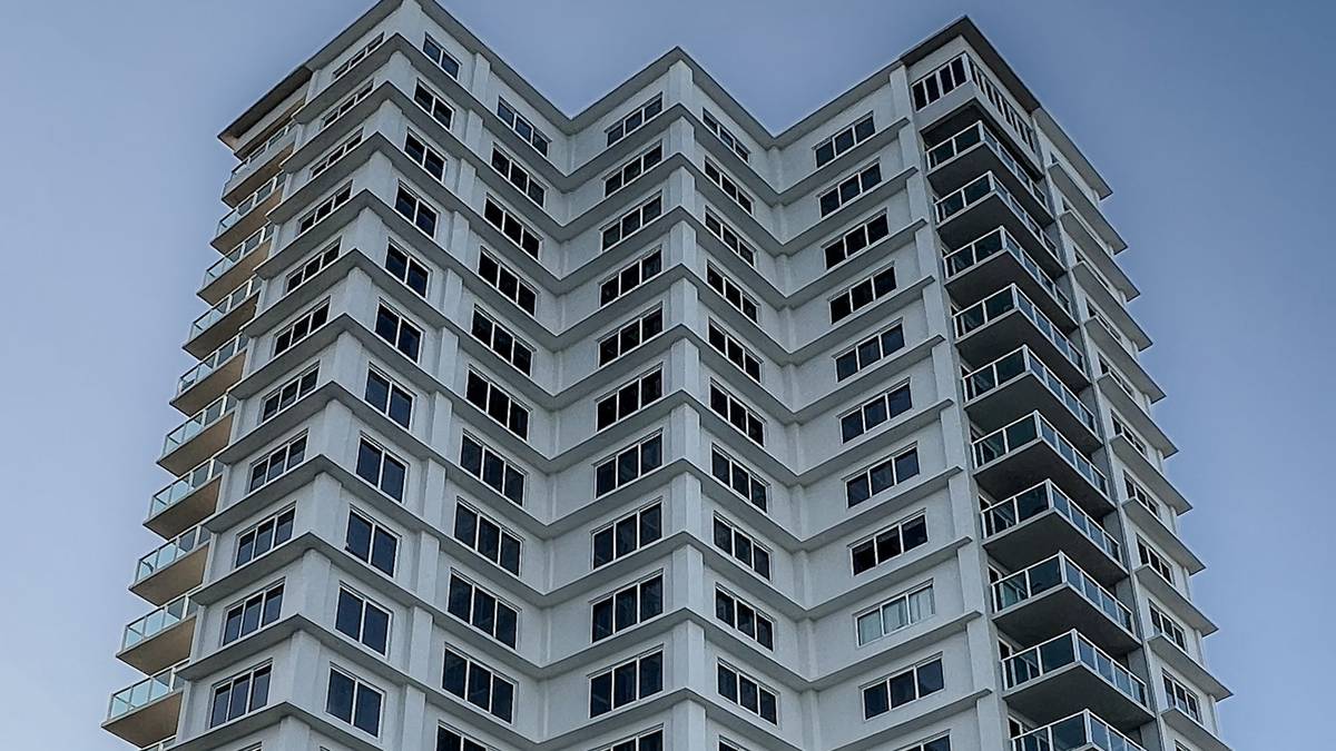Fachada de um prédio residencial alto, com sacadas de vidro e coqueiros na entrada, visto de baixo contra o céu azul