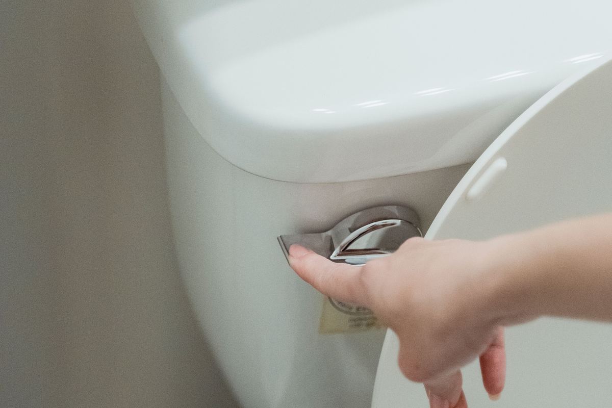 Mão pressionando a alavanca externa de acionamento da descarga de um vaso sanitário, próximo à caixa acoplada